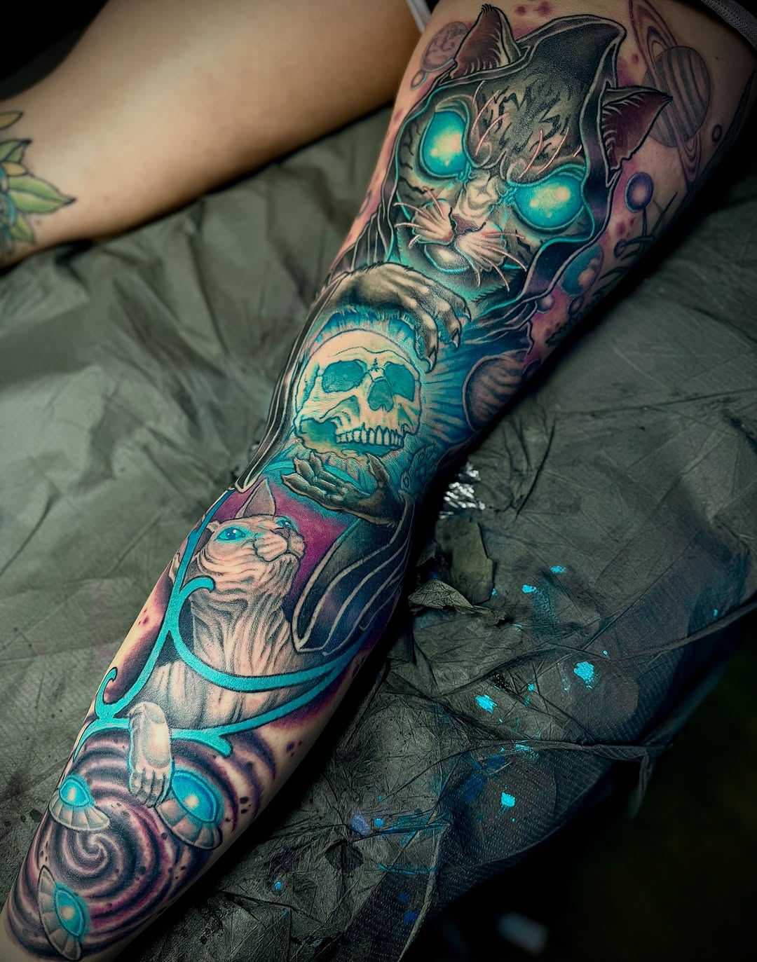Full-sleeve arm tattoo featuring a cat with glowing turquoise eyes, skull, wizard figure, and celestial elements in purple, teal, and black ink.