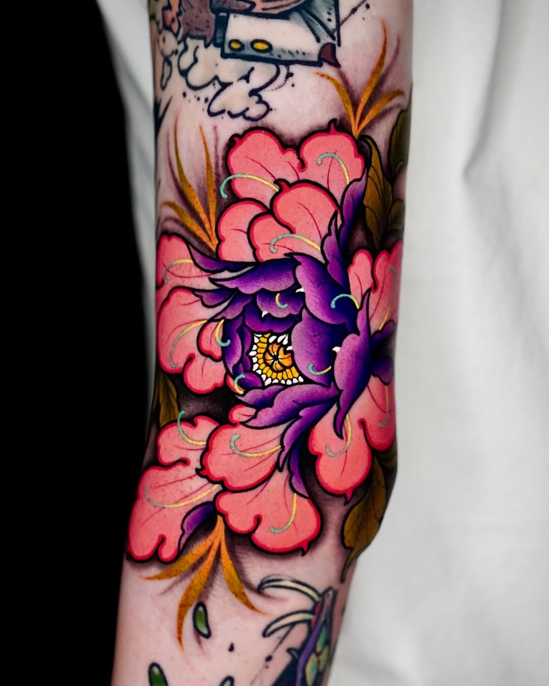 Colorful floral tattoo on forearm featuring pink and purple layered petals with yellow center and green leaves against skin.