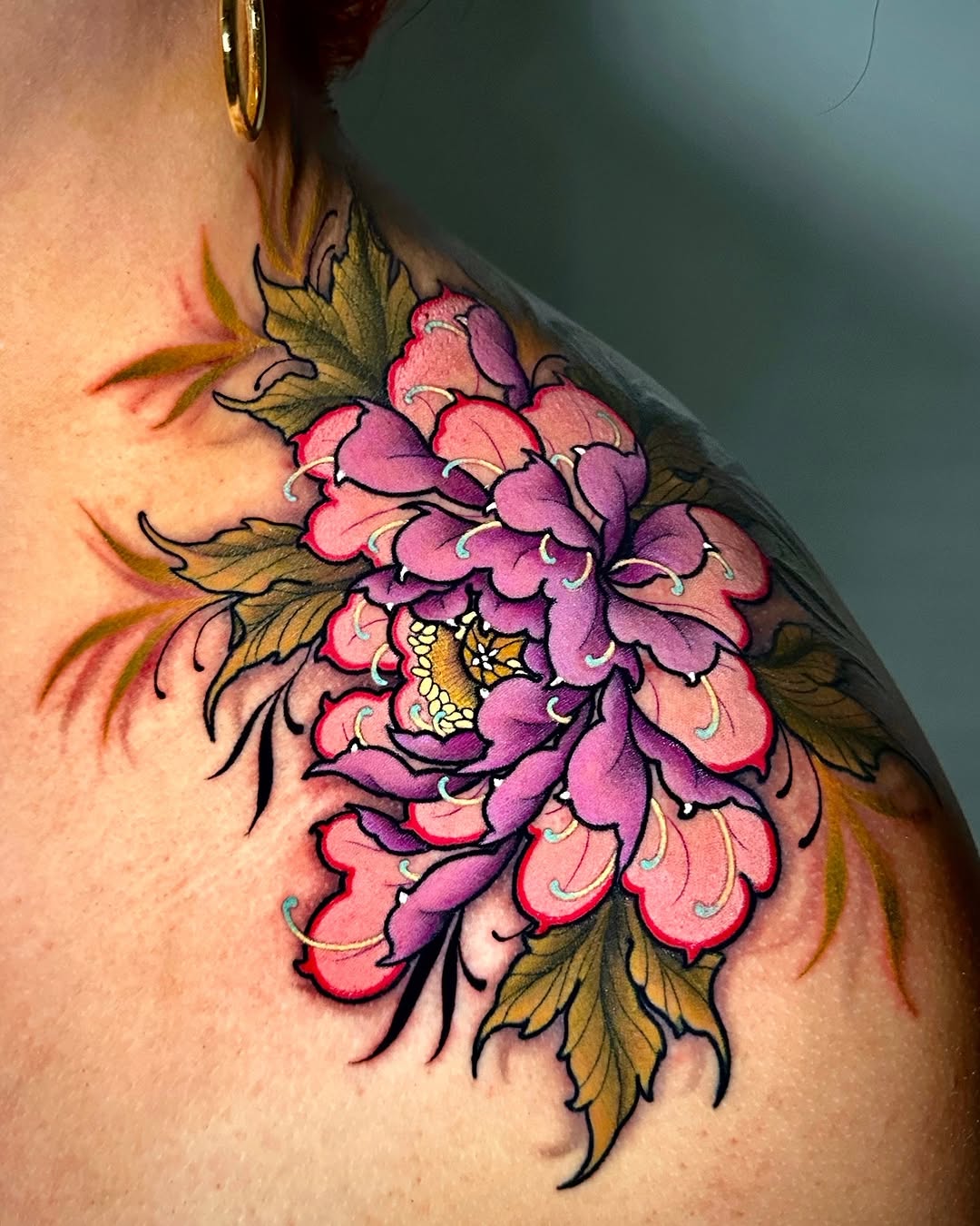 Colorful peony flower tattoo on shoulder featuring pink, purple, and gold petals with black outlines and olive-green leaves.