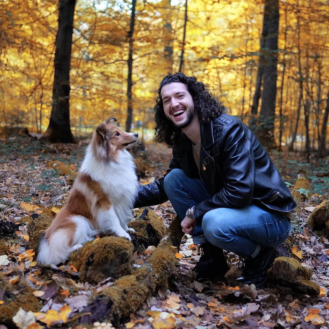 Man with long dark curly hair crouching next to a brown and white dog in an autumn forest with golden foliage.