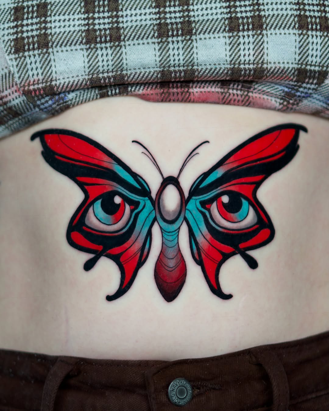 Colorful butterfly tattoo with red and turquoise wings, prominent eyes, and black outlines on lower back area.