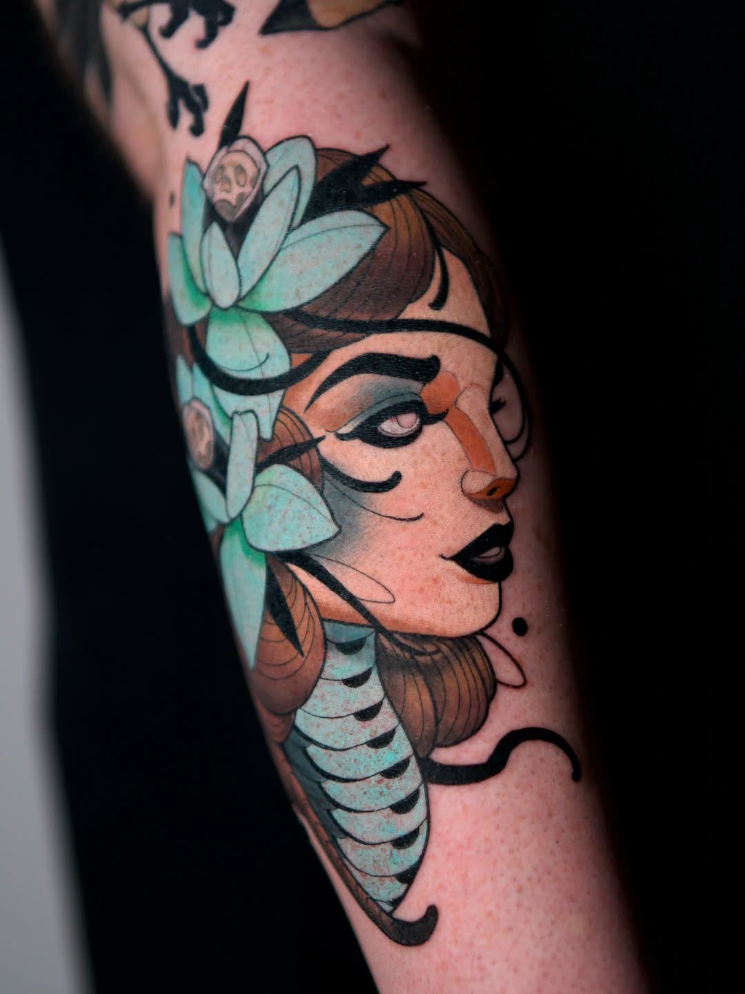Colorful tattoo on an arm featuring a woman's profile with closed eyes, flowers in her hair, and decorative scales or feathers along her neck and shoulder.