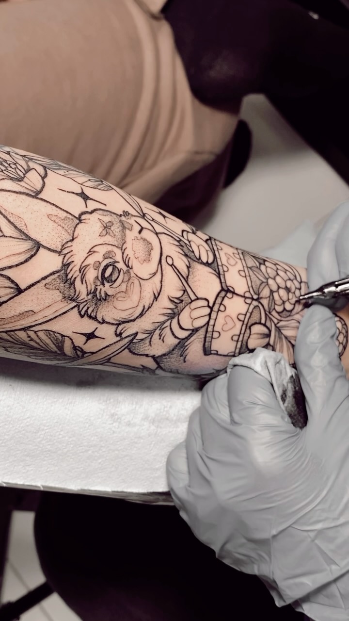 Tattoo artist inking a detailed owl with wings and floral elements on a client's forearm in black ink.
