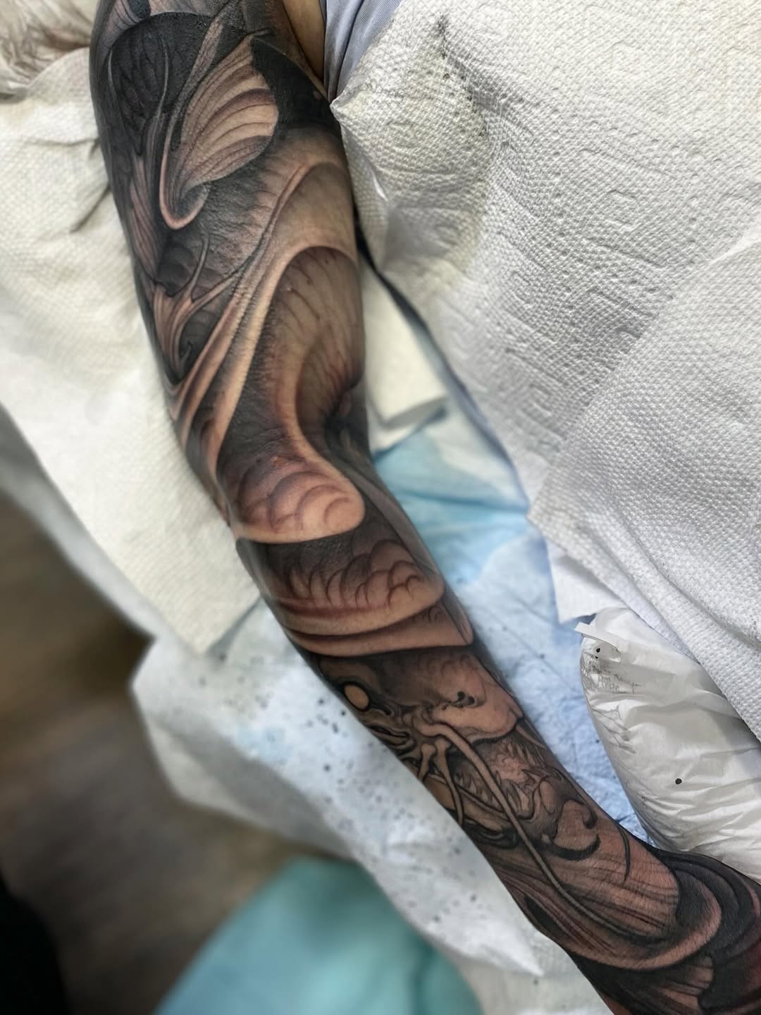 Full-color arm sleeve tattoo featuring layered abstract and organic forms in brown, black, and tan tones, displayed on a person wearing white clothing.