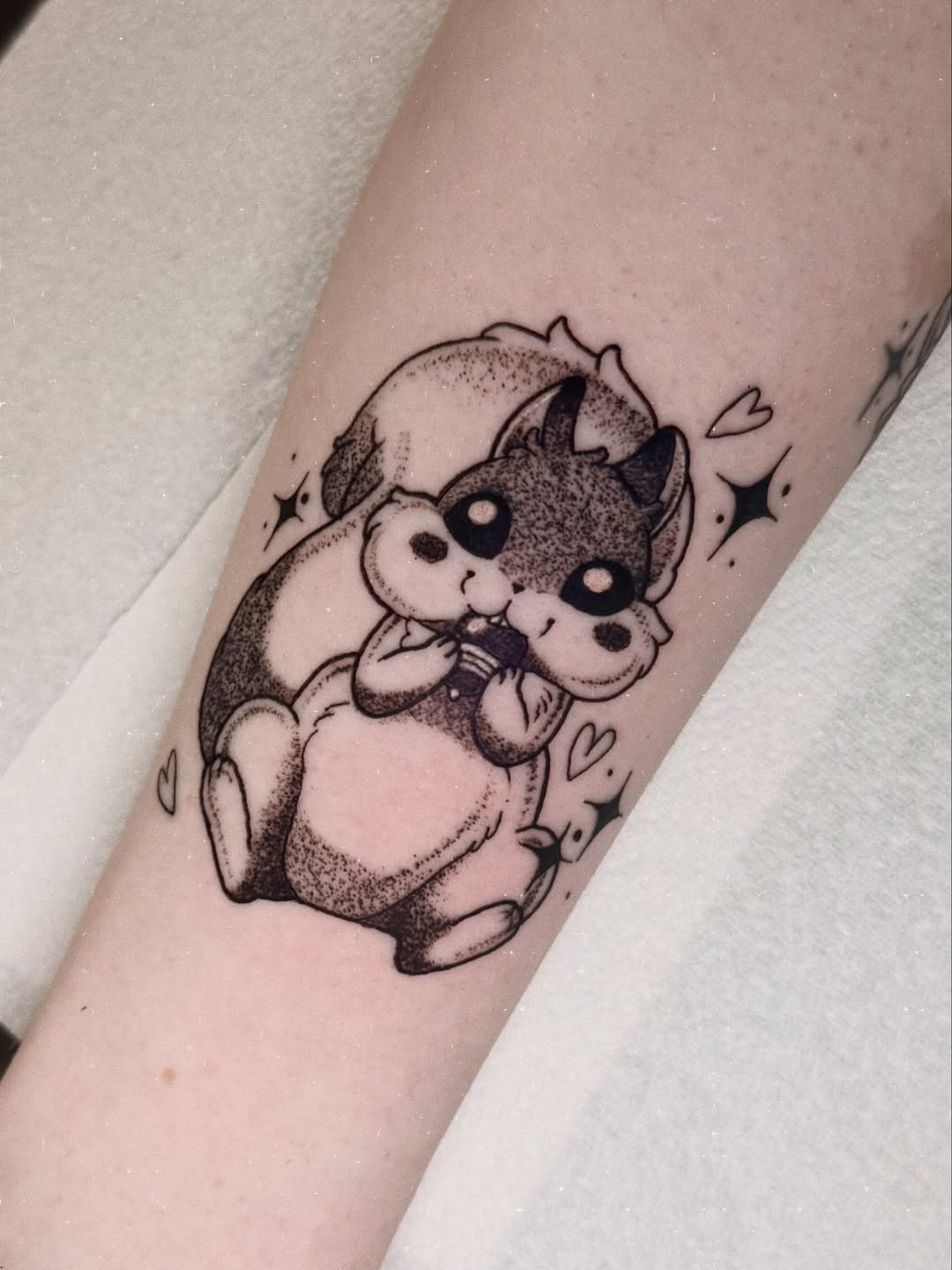Cute sitting dragon with flowers tattoo on forearm in black ink with dotted shading and small hearts and stars surrounding it.