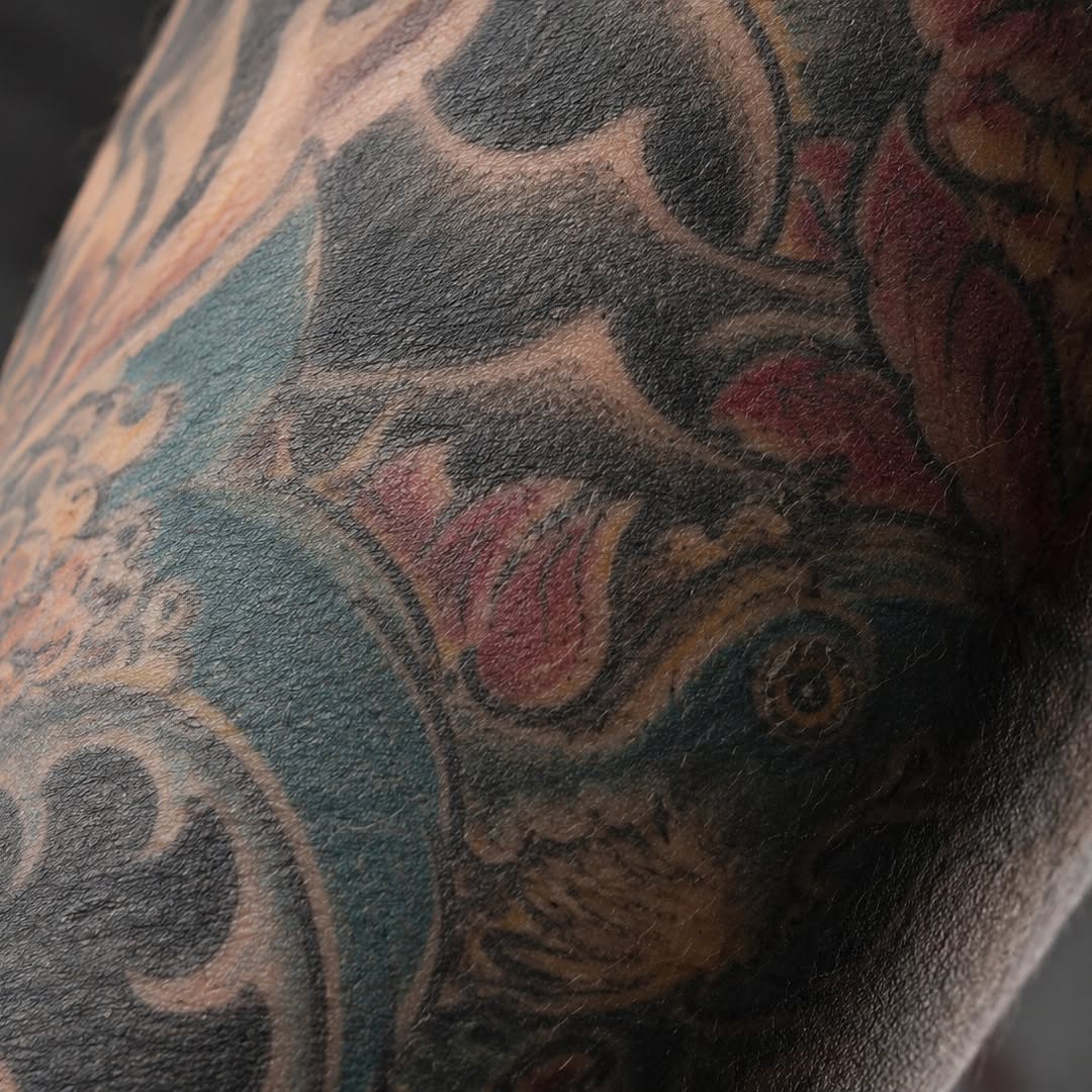 Colorful tattoo on arm featuring roses in red and burgundy tones with blue-green leaves and brown stems in detailed shading.