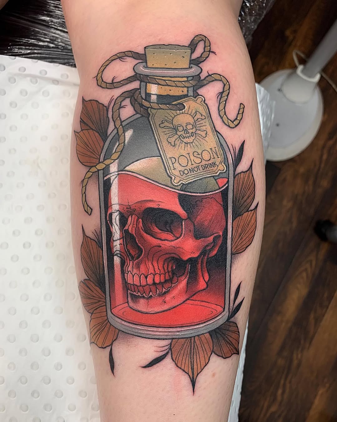 Poison bottle tattoo with red skull inside, cork stopper with rope, and brown flowers surrounding it on the calf.