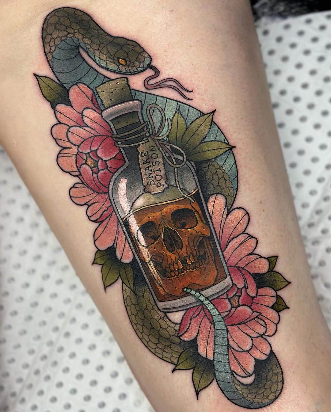 Tattoo on the arm featuring a glass bottle with a skull inside surrounded by pink flowers and coiled snakes in brown, green, and orange tones.
