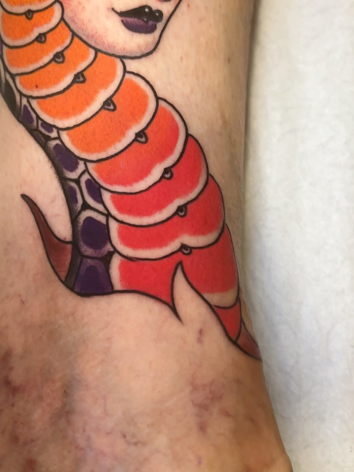 Mermaid tail tattoo in orange, red, and purple gradients with black outlines on the lower arm, featuring scaled details and a female face above.
