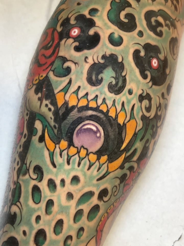 Colorful skull tattoo on an arm featuring green, black, orange, and red details with ornamental swirling patterns and circular elements throughout the design.