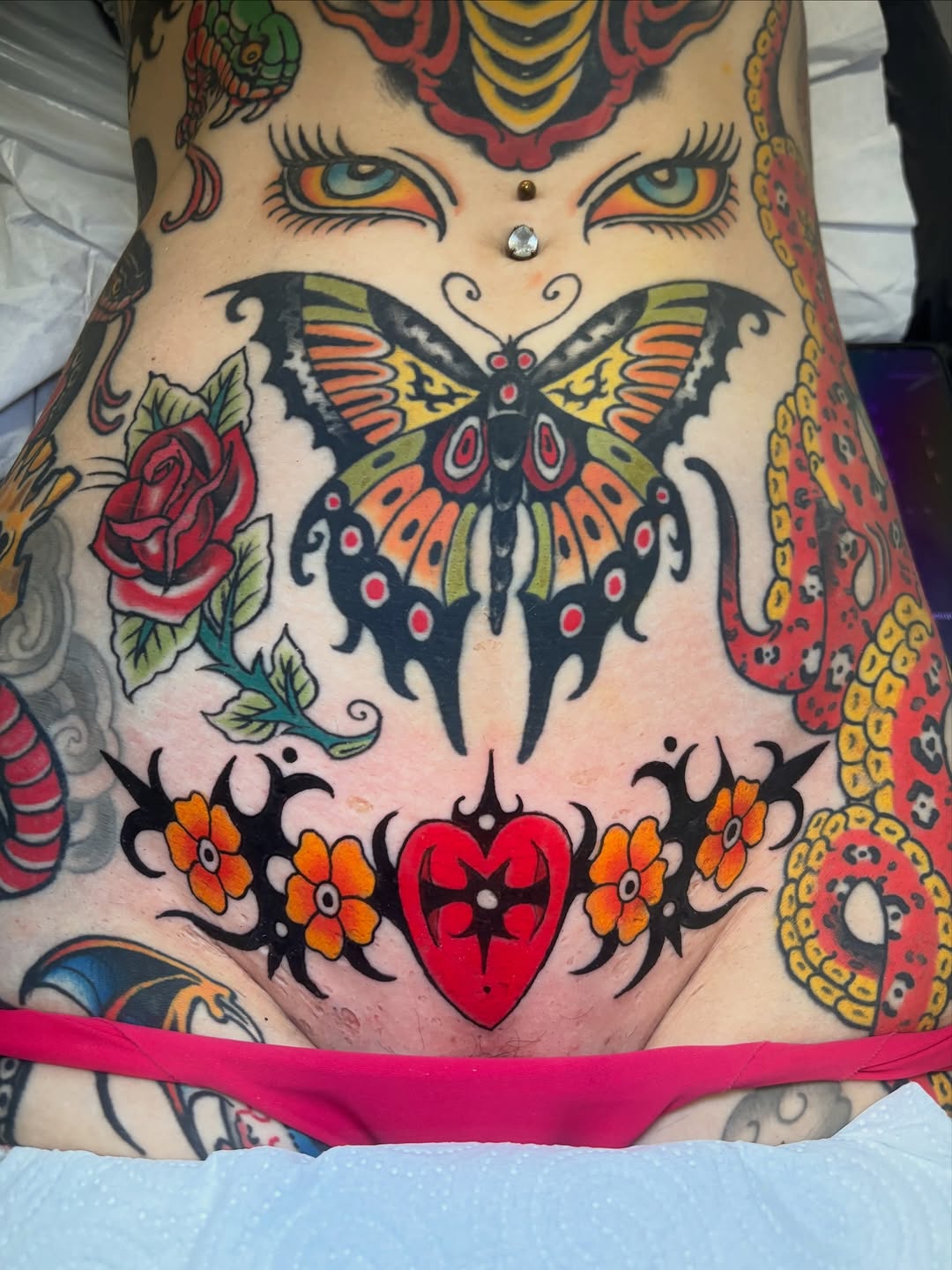 Colorful chest and abdomen tattoo featuring a face with orange eyes, butterfly, rose, heart with flowers, and ornamental patterns in red, yellow, and black.