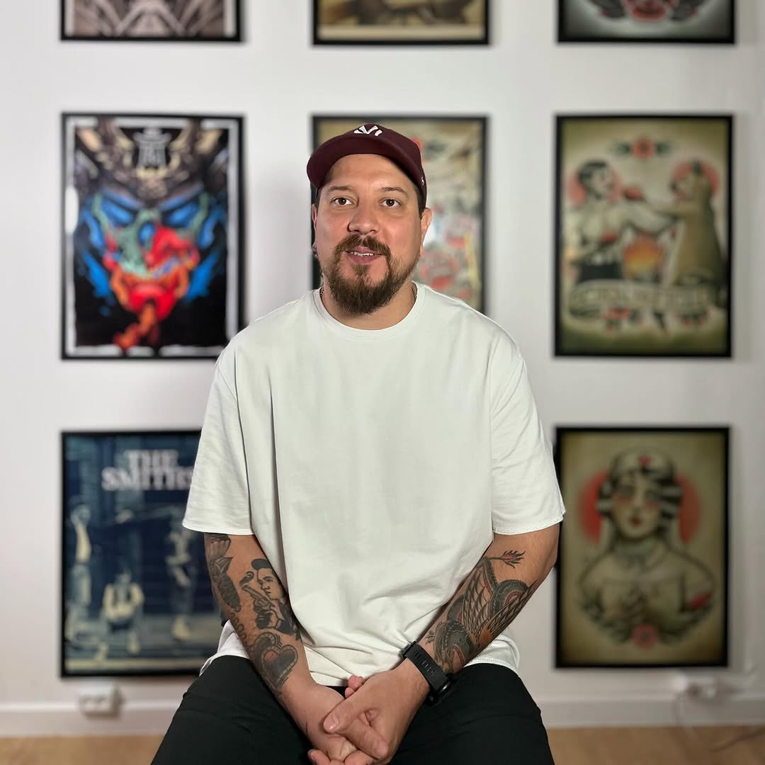 Tattooed man with facial hair wearing white t-shirt and burgundy cap, seated before framed artworks displaying colorful figure illustrations on white wall.