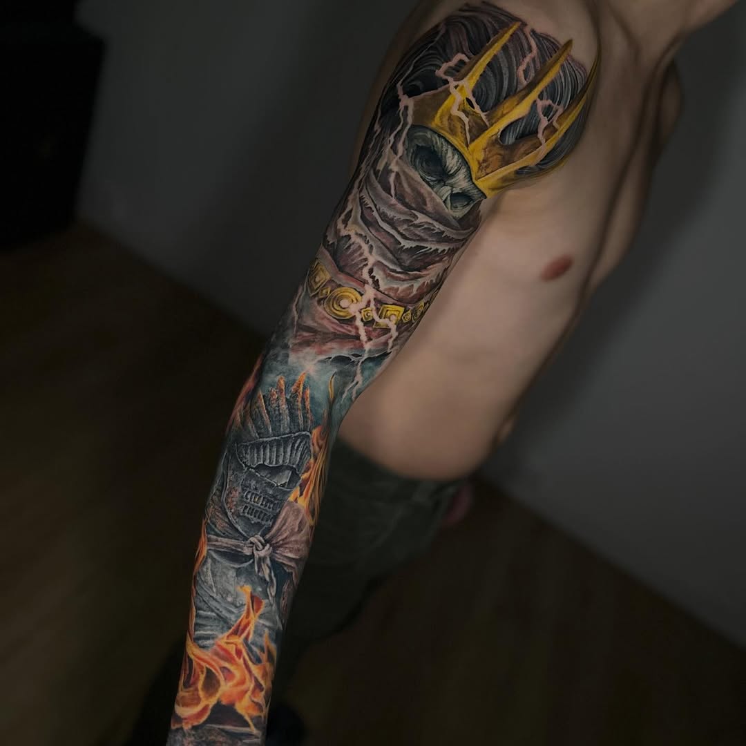 Full-sleeve arm tattoo featuring a skull with golden trident, ornate details, skeletal warrior figure, and orange flames, displayed on pale skin.