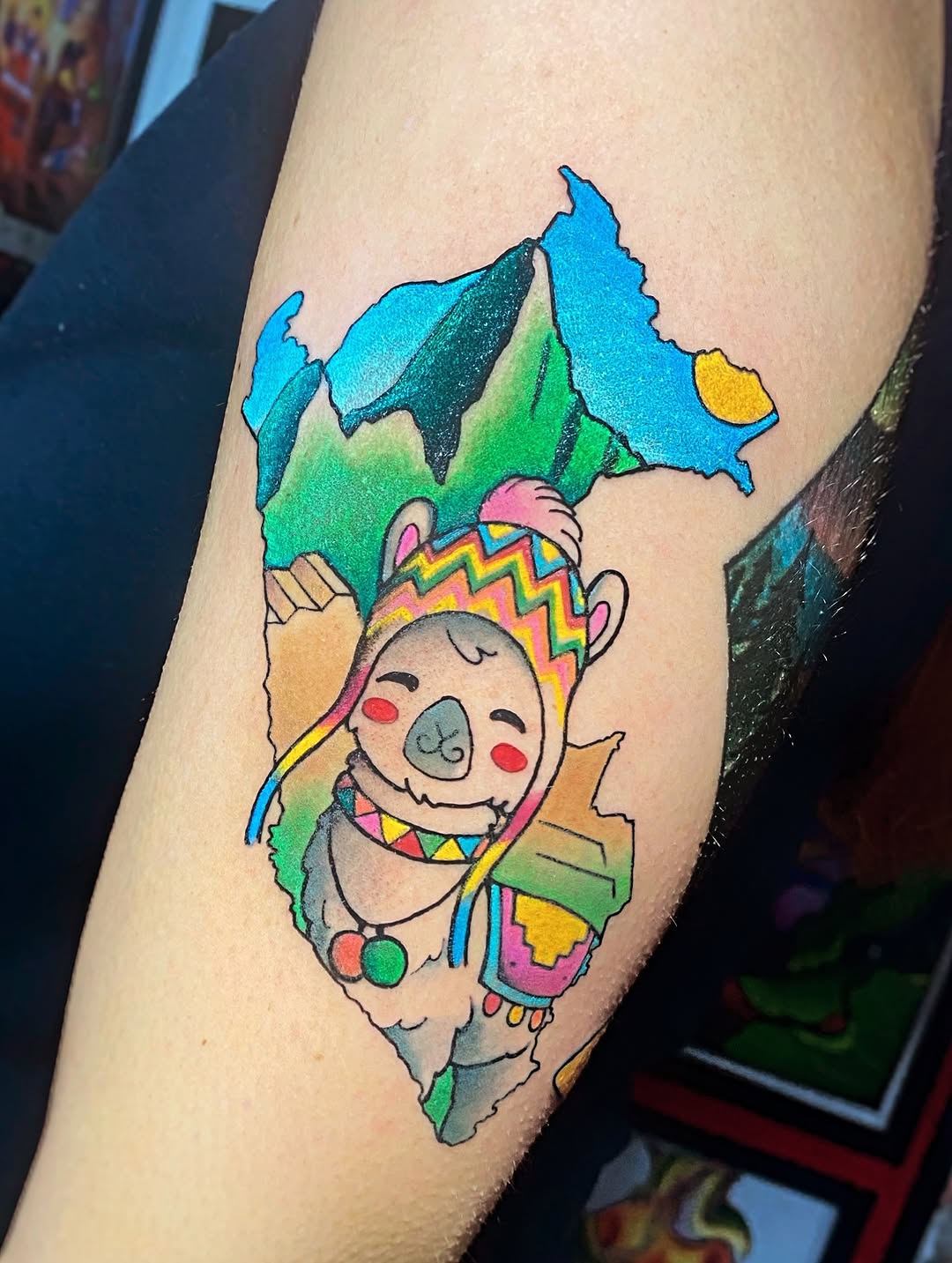 Colorful tattoo on the upper arm depicting a smiling llama wearing a decorative crown and blanket, with a map of South America in blue and green above it.