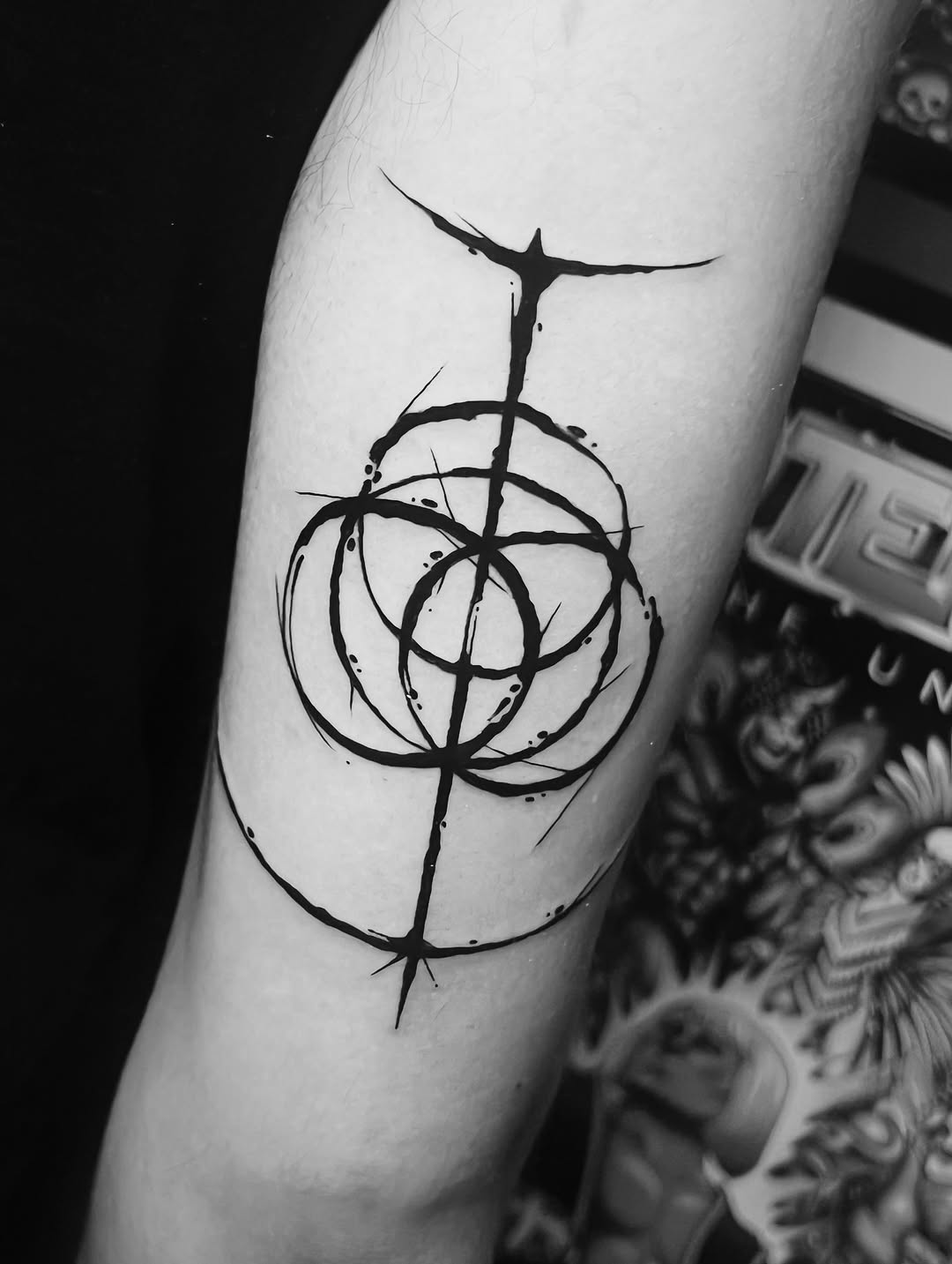 Black ink geometric tattoo on upper arm featuring concentric circles intersected by vertical and horizontal lines with radiating marks.