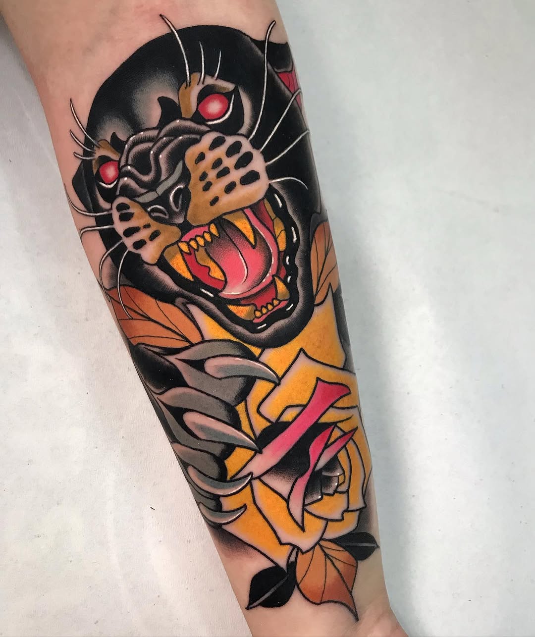 Colored tattoo on forearm featuring a roaring black cat with glowing red eyes, surrounded by orange and red flowers with pink ribbons.