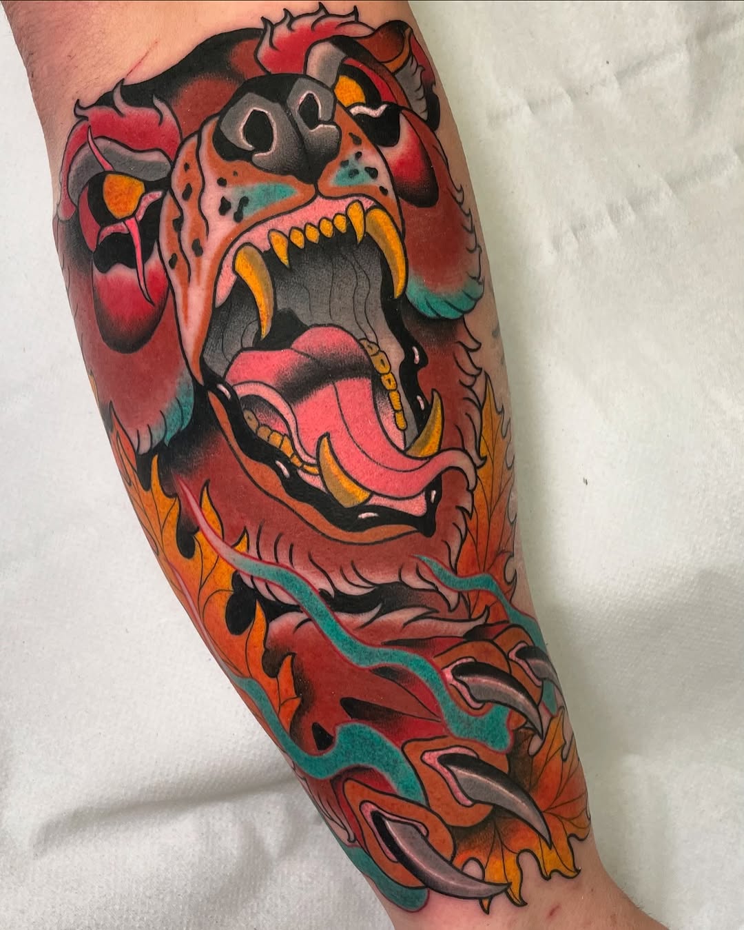 Colorful demon head tattoo with open mouth, yellow teeth, and flowing turquoise ribbon on the forearm, featuring red, orange, and black shading.