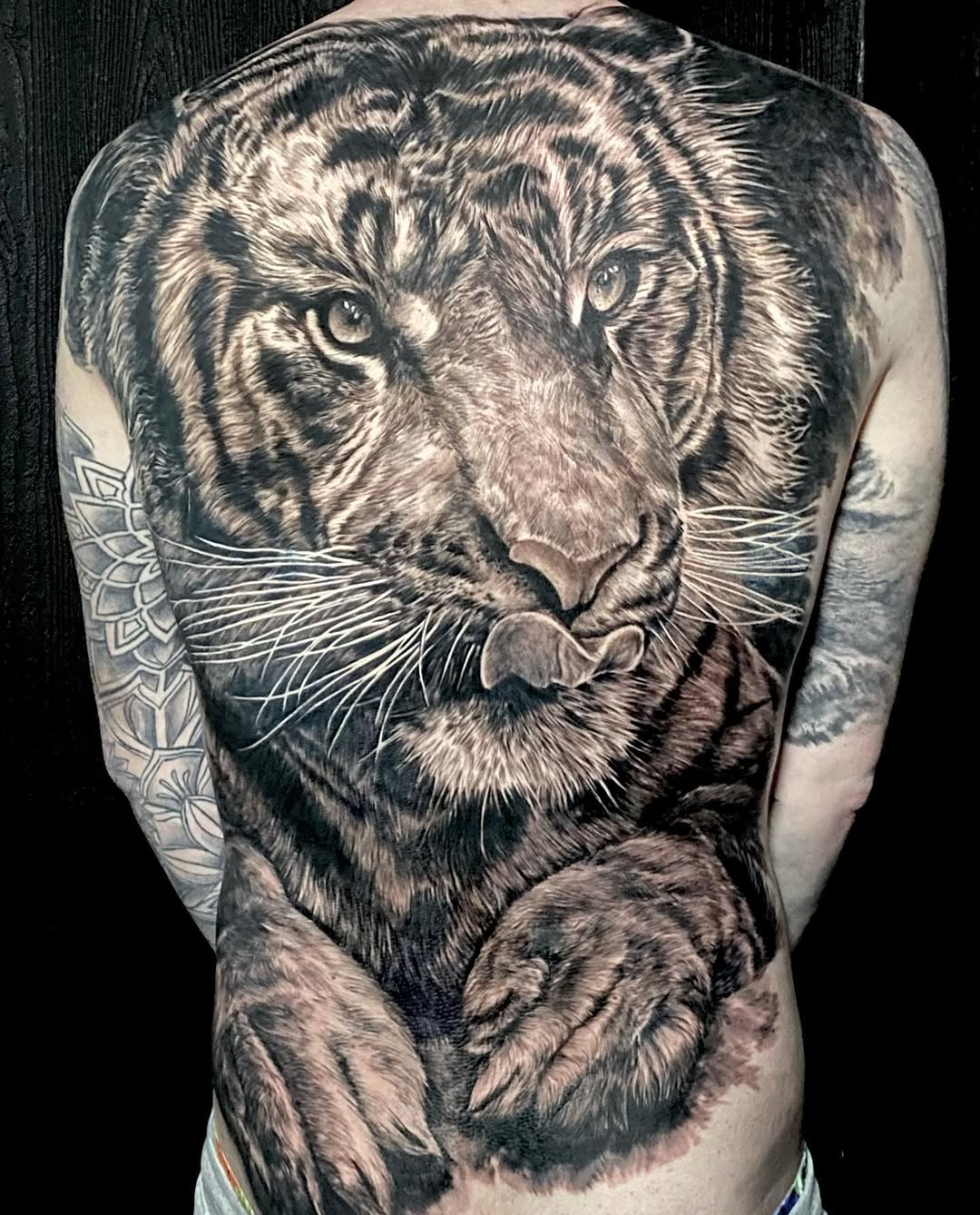Large tiger head tattoo covering the upper back and shoulders in black and grey ink, with detailed facial features and a decorative mandala pattern on the left arm.