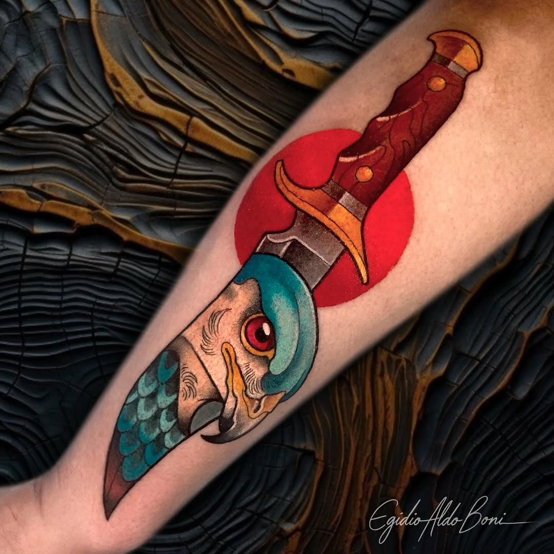Colorful tattoo on an arm featuring a dagger with a red circular background, ornate handle, and a fish-headed creature with teal and orange details below.