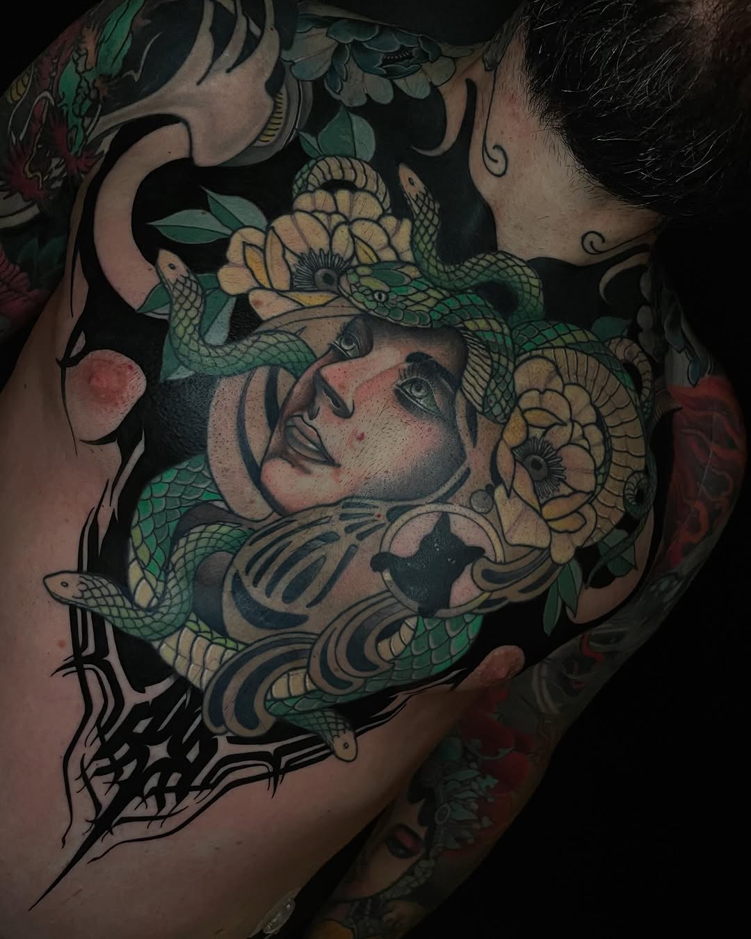 Chest tattoo featuring a woman's face adorned with green snakes and yellow flowers, with decorative black linework on the lower chest and shoulders.