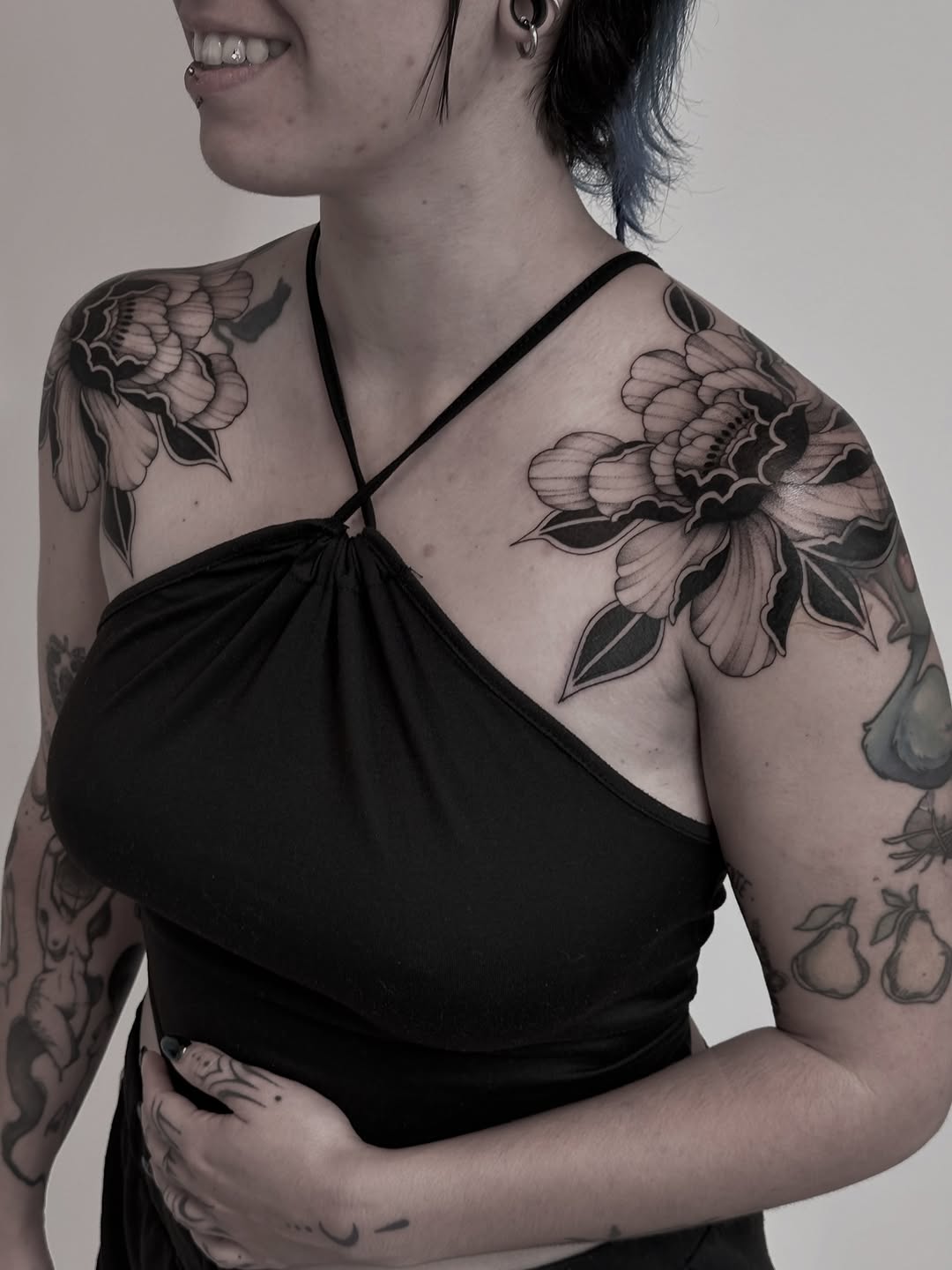 Woman displaying large-scale floral shoulder and upper arm tattoos in black ink on pale skin, wearing a black halter top.