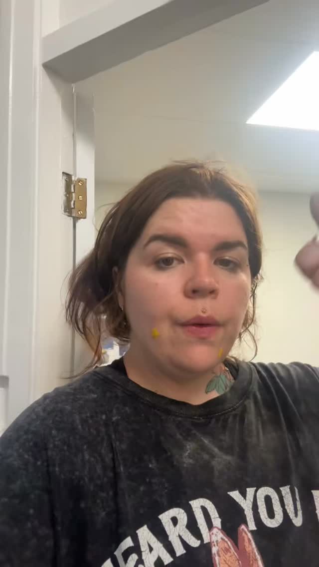 Person wearing dark graphic t-shirt displaying a small yellow dot tattoo on their chin.