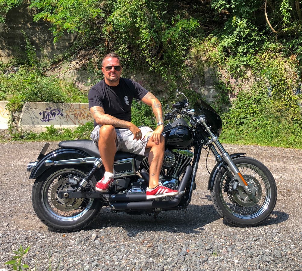 Man with a sleeve tattoo featuring dark ink designs on his right arm, posing on a motorcycle outdoors.
