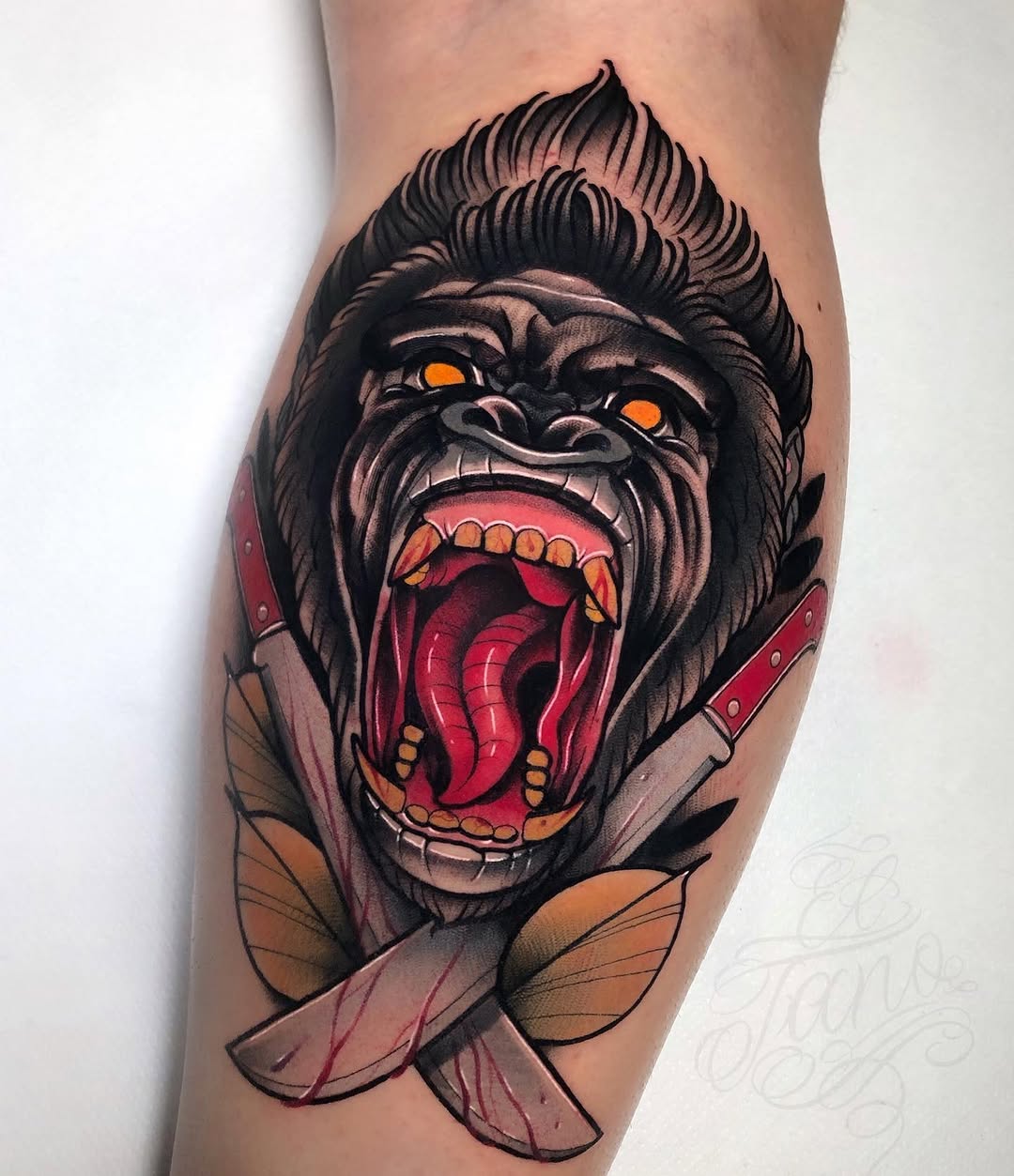 Angry ape head with glowing orange eyes and open red mouth holding a knife, tattooed on a calf with brown and red coloring.