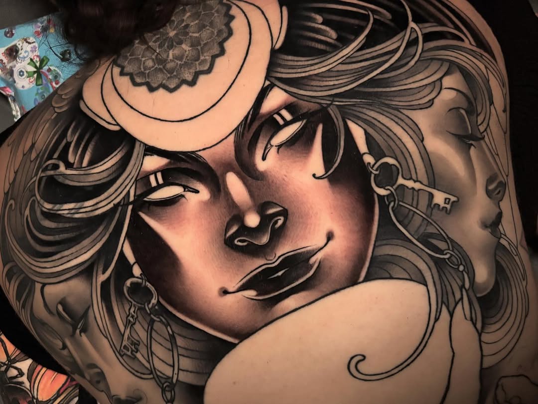 Two women's faces with detailed shading and line work in black ink on skin, featuring flowing hair, jewelry, and a mandala design on the shoulder.