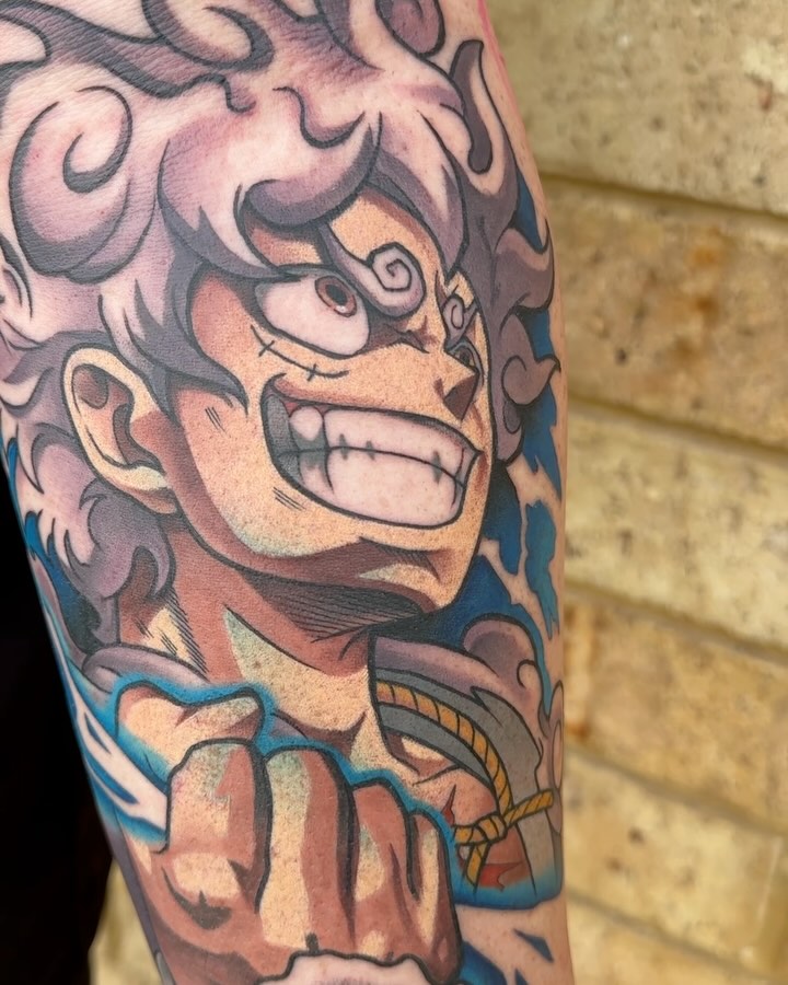 Colorful demon face tattoo on upper arm with ornamental scrollwork, featuring red, blue, and gold accents on pale skin against a brick wall.