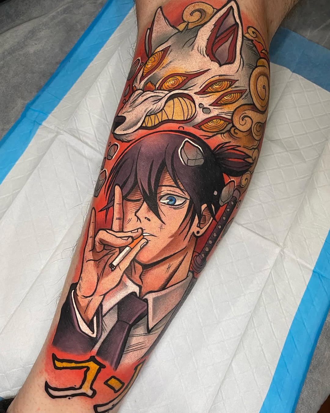 Colorful leg tattoo featuring a human face smoking a cigarette with a demon mask above, rendered in red, orange, gold, and black tones.