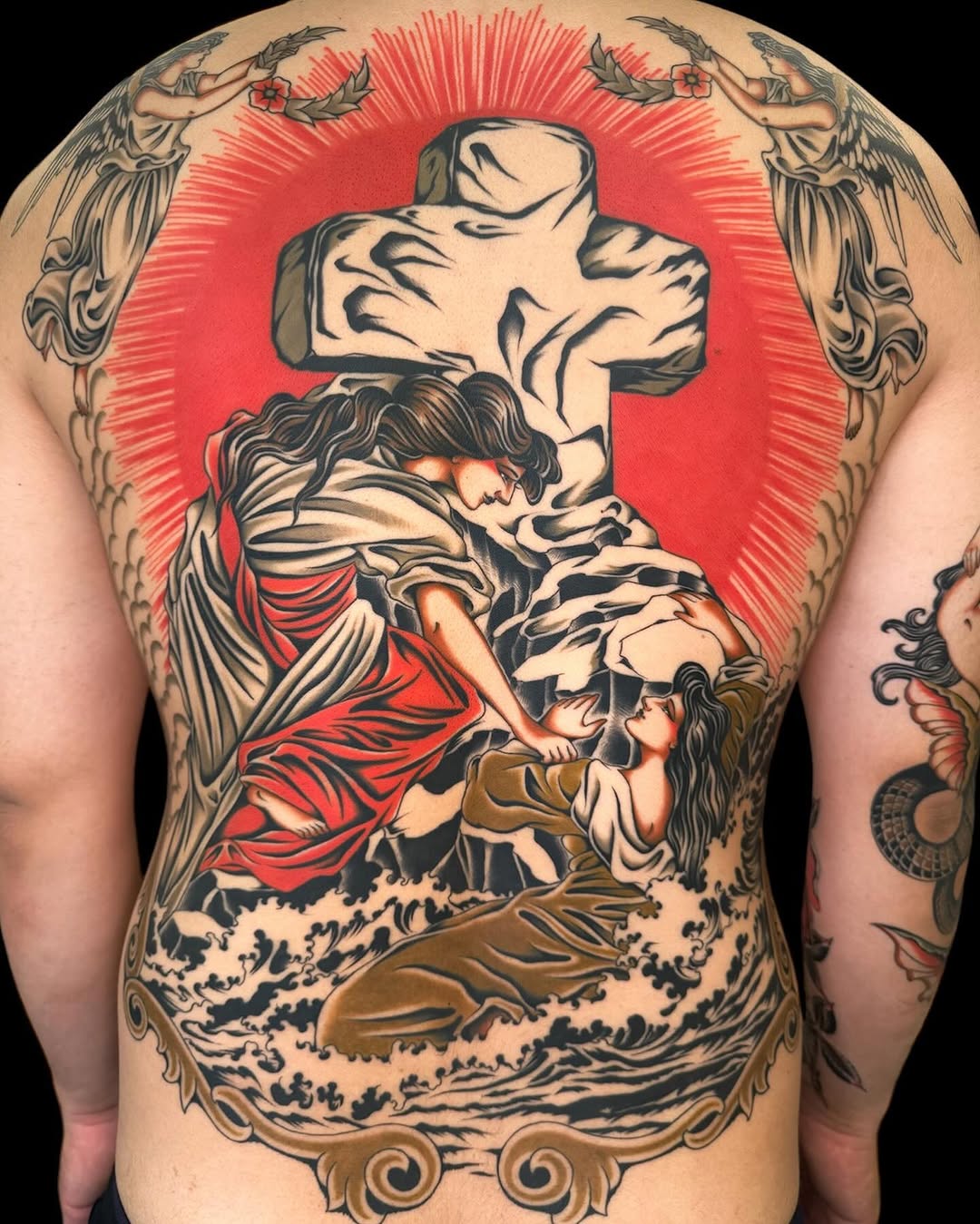 Large back tattoo featuring a glowing red cross, angels with wings, a robed figure, cherubs, ocean waves, and ornamental scrollwork in black, red, and brown ink.