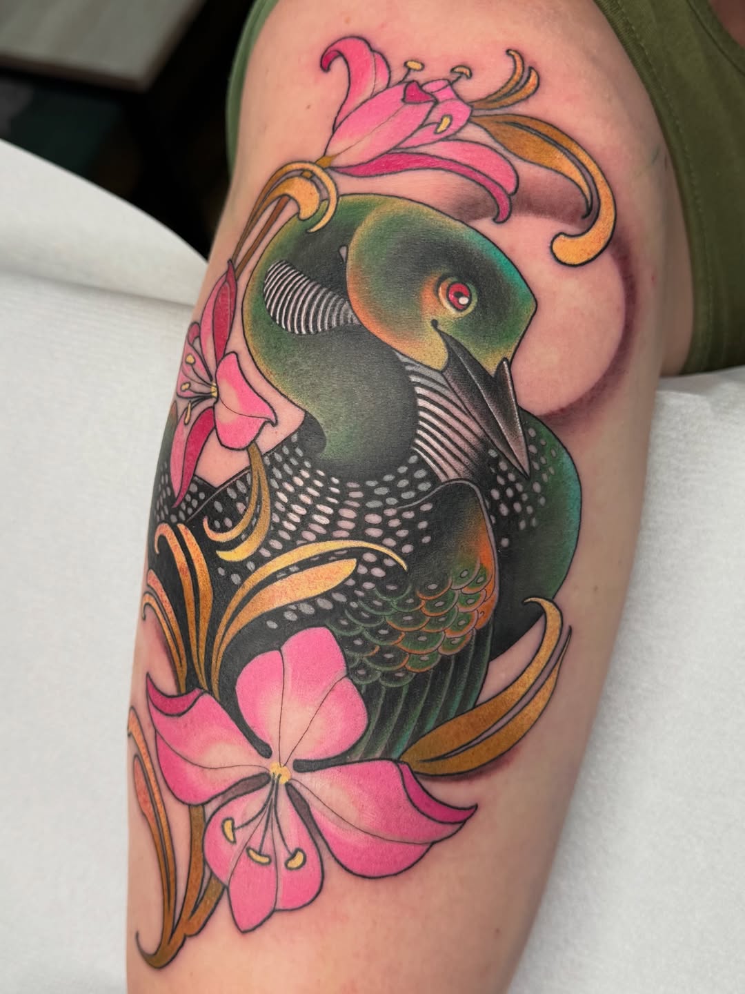 Colorful fish tattoo on upper arm featuring a green and black fish with pink flowers and gold scrollwork accents.