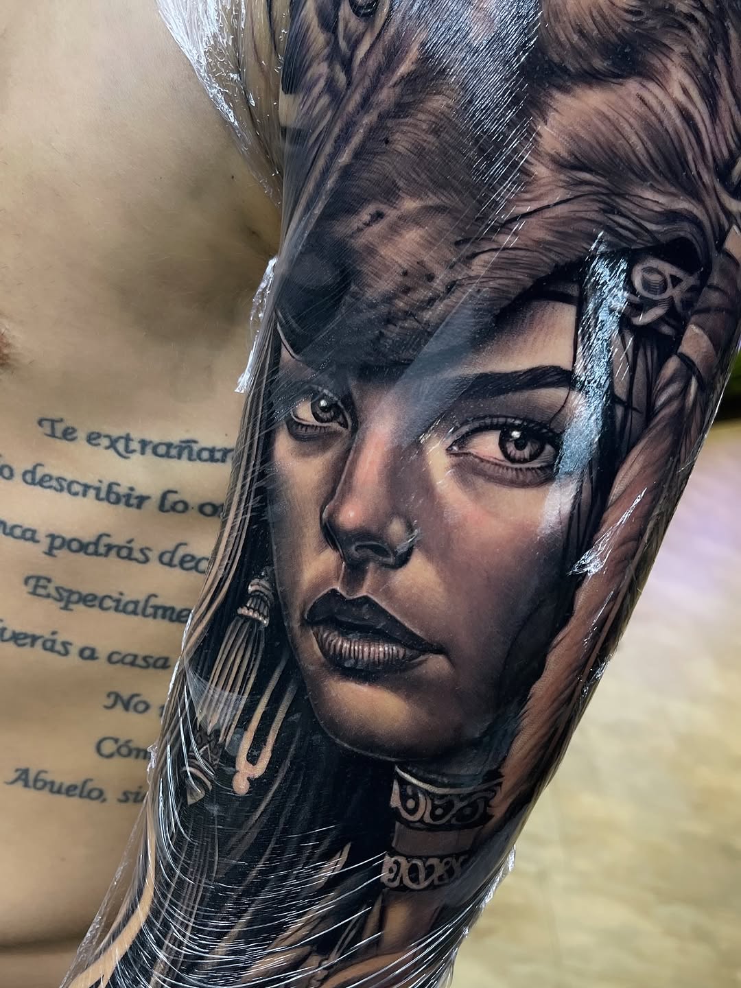Portrait tattoo of a woman's face with long hair, decorative neck ornaments, and Spanish text on the upper arm, done in black ink on skin.