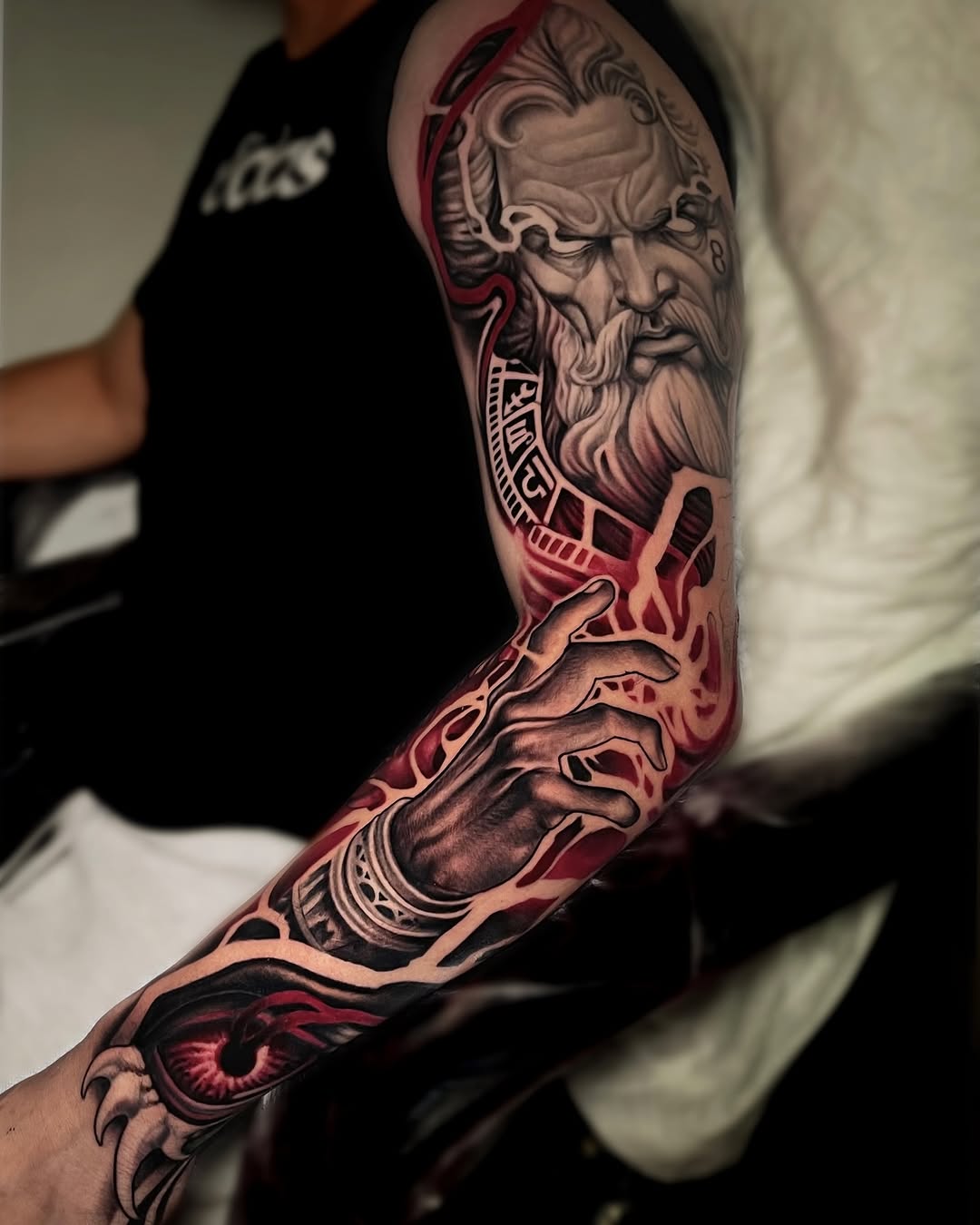 Full-sleeve arm tattoo featuring a bearded male face, ornamental patterns, and a red eye design in grayscale and red ink.