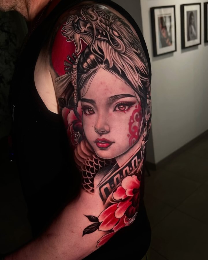 Colorful shoulder tattoo featuring a woman's face with ornate headpiece, dragon and phoenix imagery, red peonies, and decorative patterns in red, pink, and black ink.