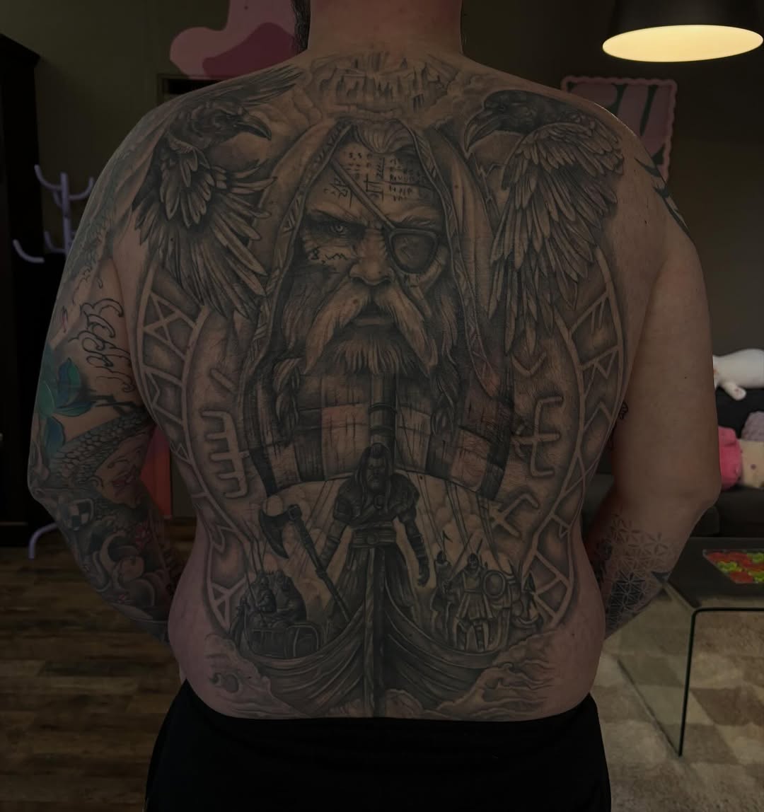 Large back tattoo featuring a bearded man with an eye patch and winged figures, surrounded by ornate geometric patterns and runic symbols in black and gray ink.