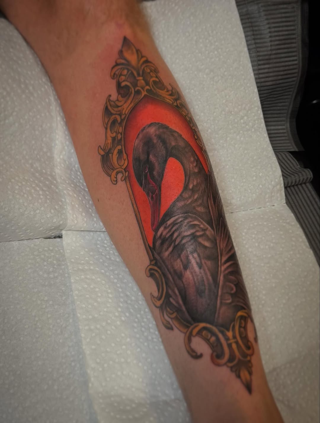 Heart tattoo with dark figure silhouette and bright red background, framed by ornate gold scrollwork, on the forearm.