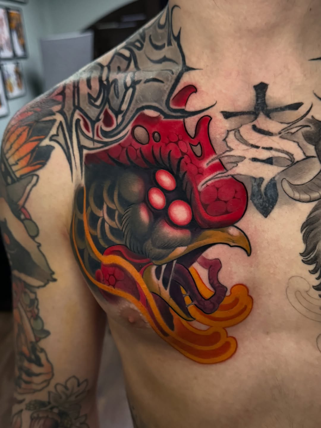 Colorful demon or oni face tattoo on chest featuring red skin, large eyes, prominent teeth, orange flames, and black line work accents.
