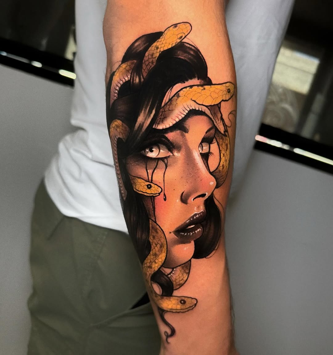 A woman's portrait with snakes intertwined around her head, rendered in orange and black ink on a forearm.