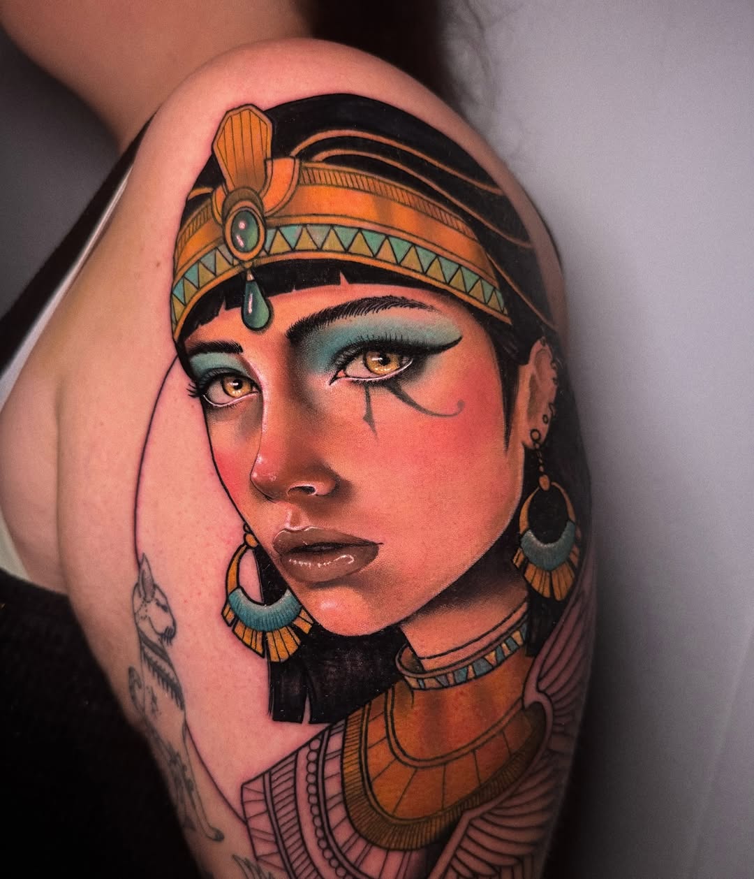 Colorful shoulder tattoo depicting an Egyptian-inspired female figure with ornate headdress, jewelry, and Egyptian Eye of Horus makeup in orange, teal, and gold tones.