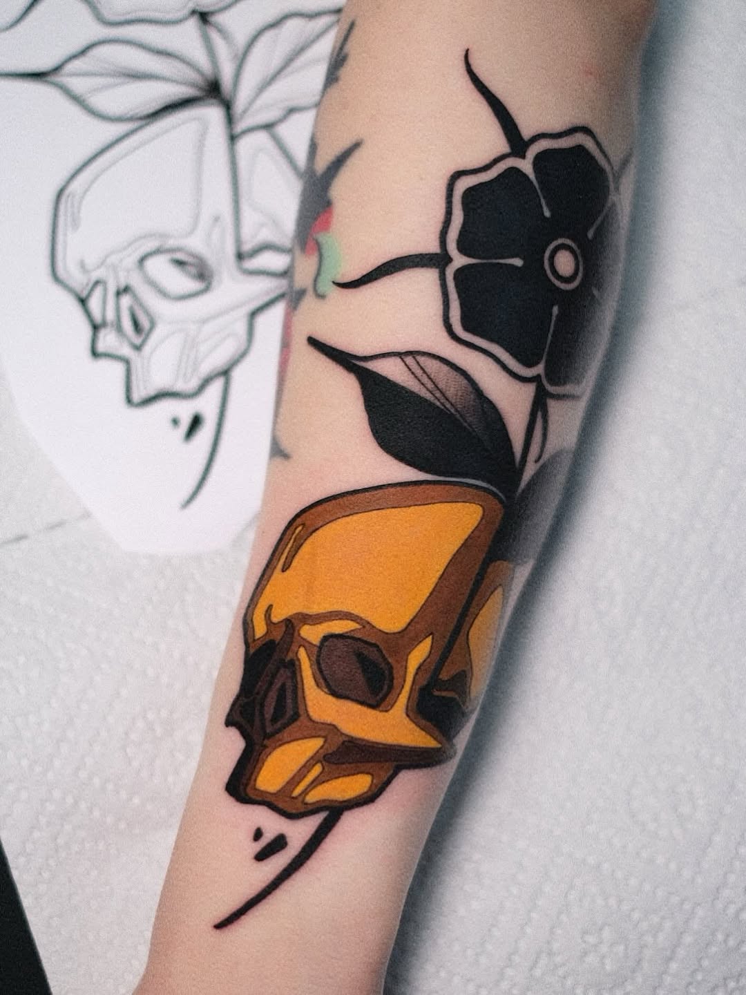 Vertical arm tattoo featuring a black flower with a pink center, black leaves, and an orange/yellow geometric shape with brown shading and decorative lines.