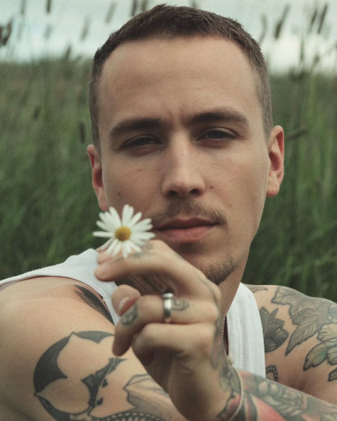 Tattooed man holding a white daisy flower, displaying leaf and botanical designs on both arms in green and black ink.