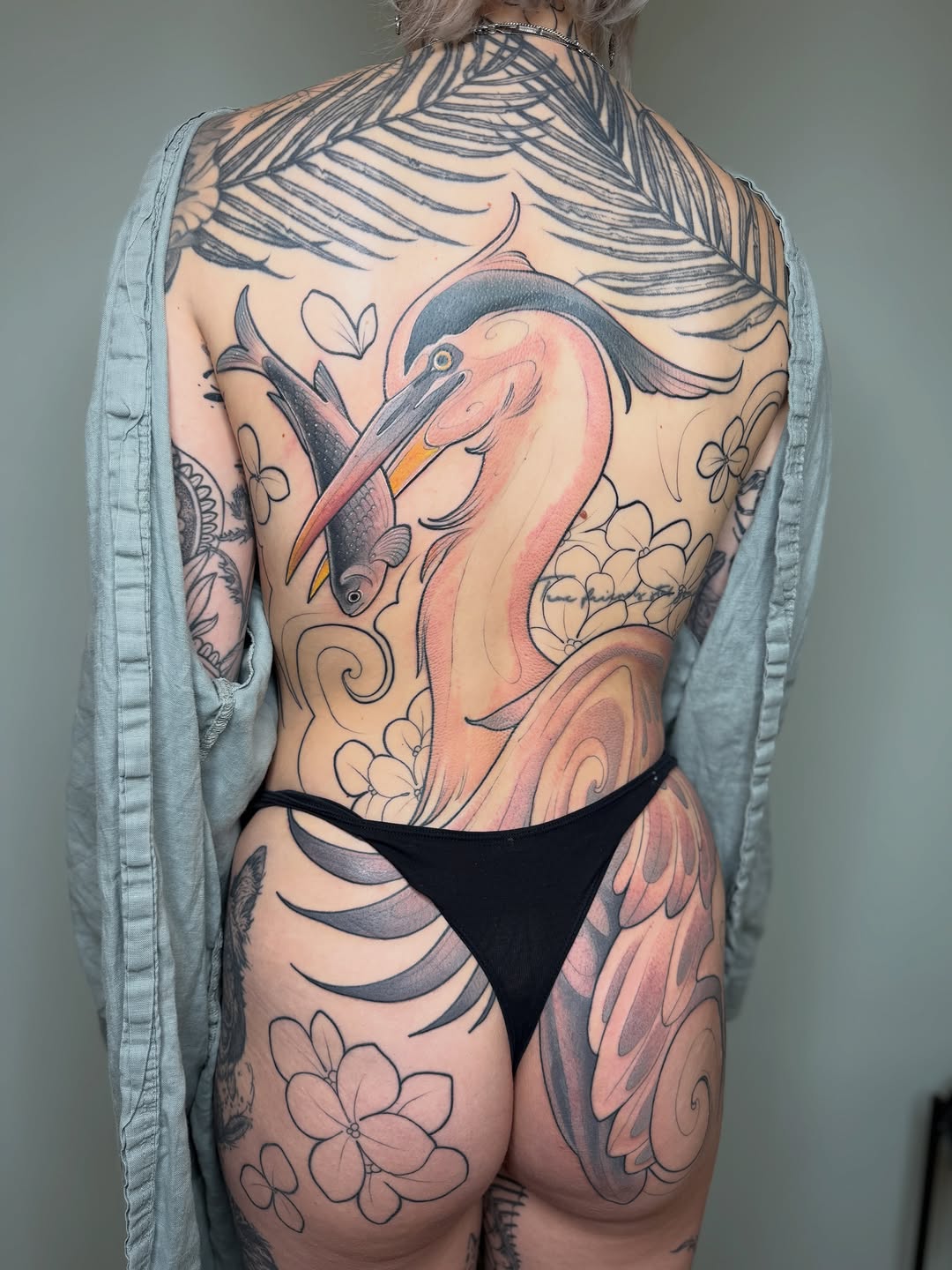 Back tattoo featuring a pink flamingo with extended wings, surrounded by feather designs, floral elements, and decorative swirls in black and colored ink.