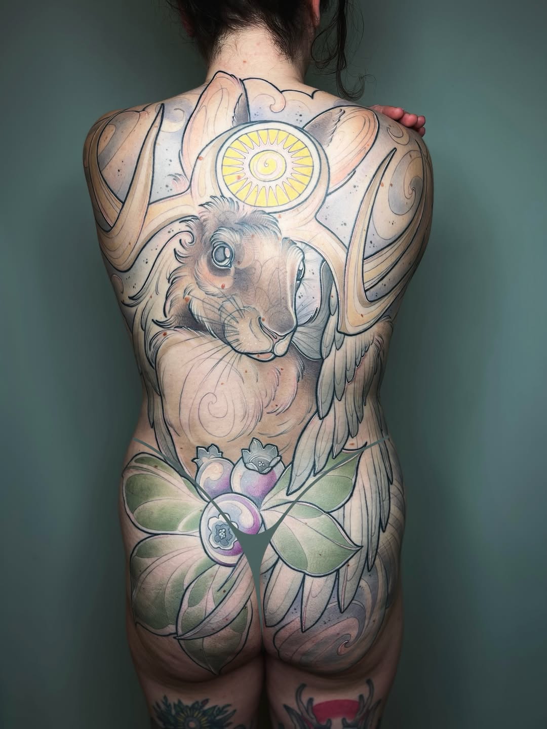 Back piece tattoo featuring a winged lion with blue eyes and a yellow sun above it, surrounded by ornamental designs and purple flowers with green leaves.