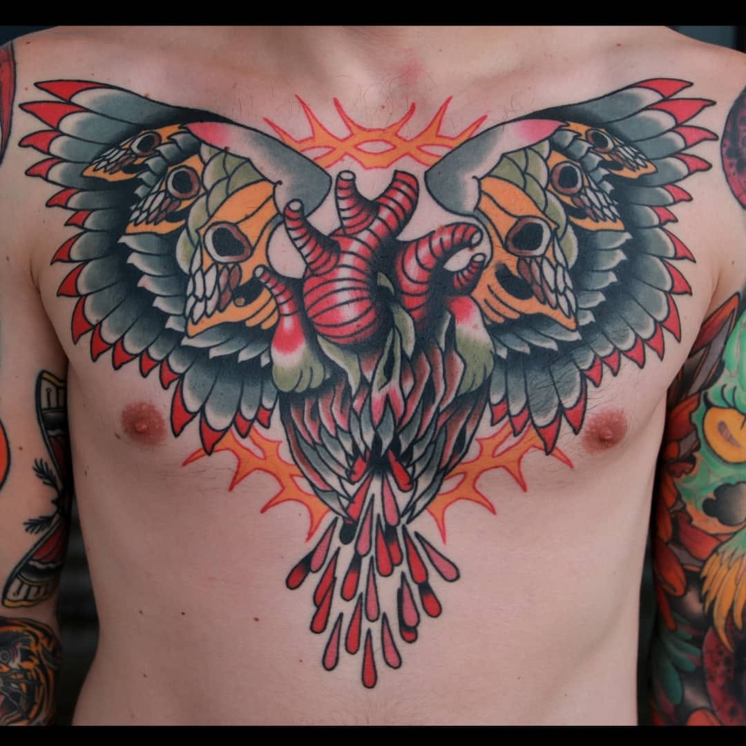 Colorful chest tattoo featuring a winged skull with spread wings, orange and red flames, and intricate feather details in red, orange, black, and grey.