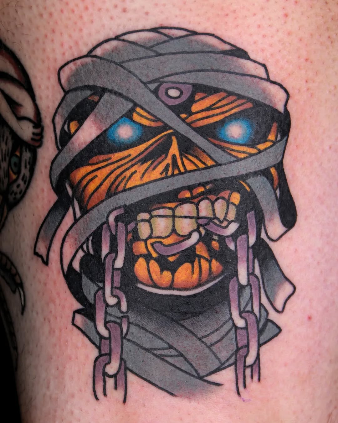 Mummy head tattoo with glowing blue eyes, orange bandage patterns, bared teeth, and dangling strips on pale skin.