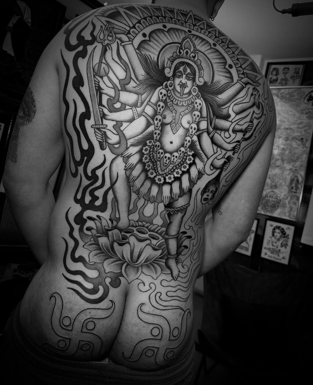 Large back tattoo featuring a multi-armed deity figure with ornate crown and jewelry, surrounded by decorative floral and serpentine motifs in black ink.