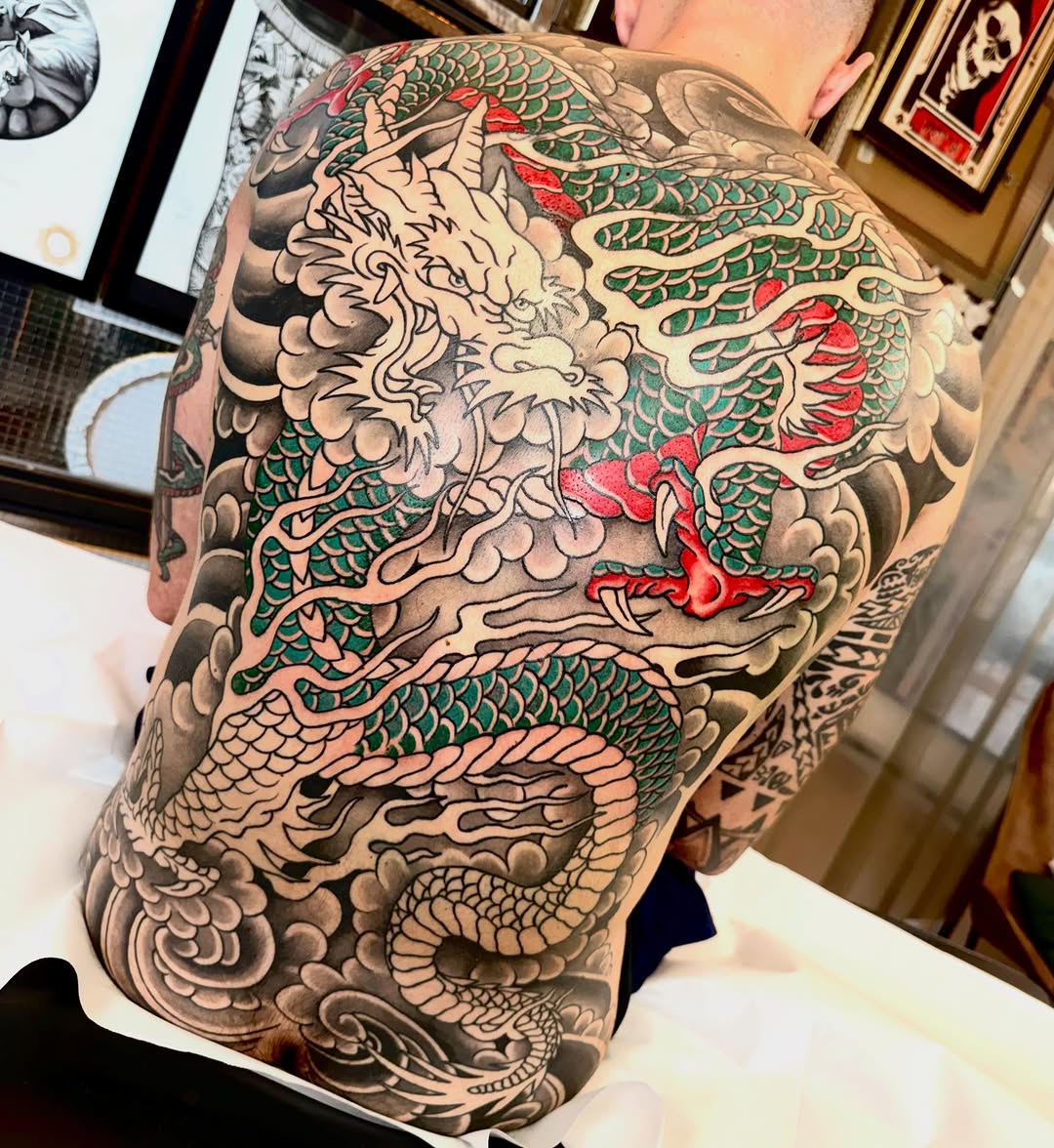 Dragon and mythical creature tattoo on shoulder and arm with green scales, red accents, and grey shading throughout the design.