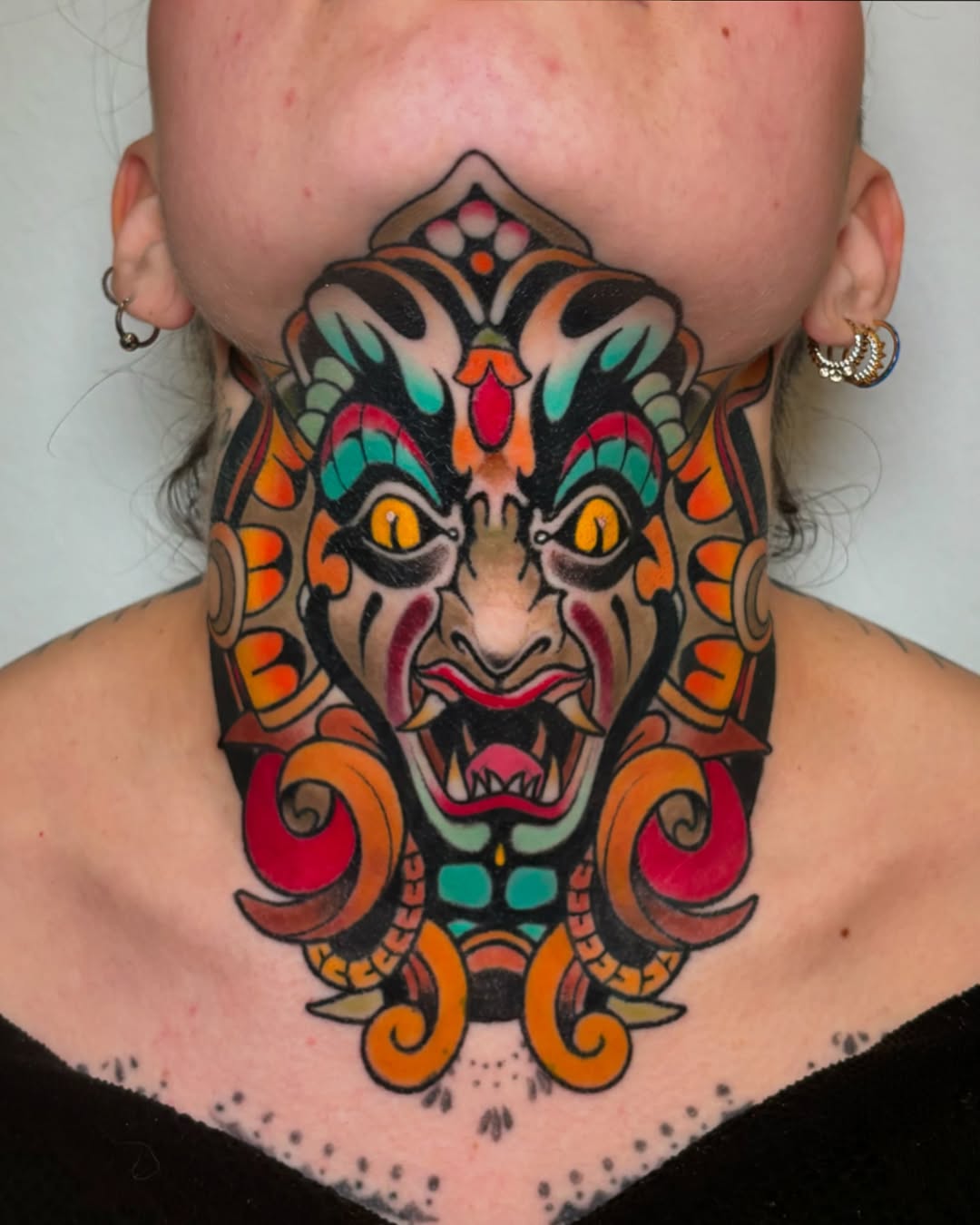 Colorful demon mask tattoo on neck and chest featuring golden eyes, red mouth, orange scrollwork, and turquoise accents with symmetrical design.